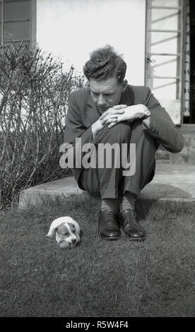 1950 s, historischen, einen geeigneten Mann sitzt auf einem Schritt in einem Garten neben einem kleinen Jack Russell Terrier Hund. Der Jack Russell Terrier wurde ursprünglich als working Terrier zu jagen Füchse gezüchtet und ist eine lebendige, unabhängige und kluge kleinen Hund. Stockfoto