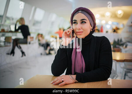 Attraktive junge arabische Frau sitzen an einem Tisch in Cafe. Sie spricht am Telefon. Modell lächelt. Sie trägt Hijab Stockfoto