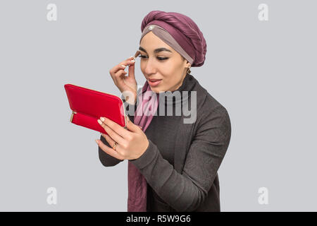 Schöne und attraktive junge arabische Frau tun Make-up. Malen Sie Ihre Augen und schauen Sie in den Spiegel. Auf grauem Hintergrund Stockfoto