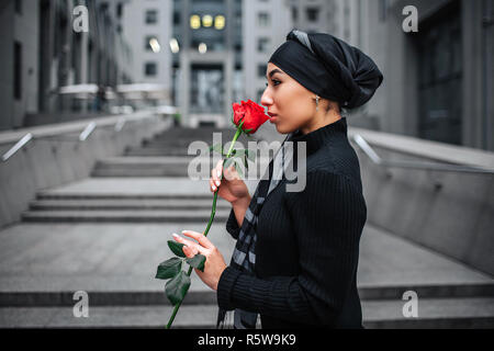 Die Side Shot der jungen arabischen Frau stehen auf Schritte und rote Rose in den Händen halten. Sie riecht. Das Modell trägt schwarz Hijab Stockfoto