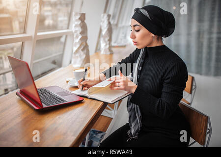 Konzentrierten arabischen Frau im schwarzen Kleid und Hijab. Sie sitzt in der Seite am Tisch und Weiß in Notebook. Laptop und Tasse Kaffee auf Tisch Stockfoto