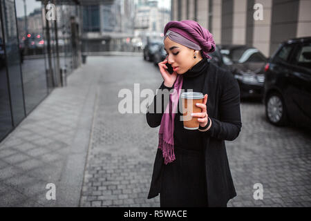 Besetzt und ernsthafte junge arabische Frau im Freien stehen und nach unten schauen. Sie spricht am Telefon und halten Sie die Tasse trinken Stockfoto