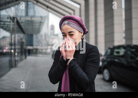 Kranke junge arabische Frau im Freien stehen kann. Sie Niest in Weiß Serviette. Das Modell trägt schwarze Kleidung und Violett hijab Stockfoto