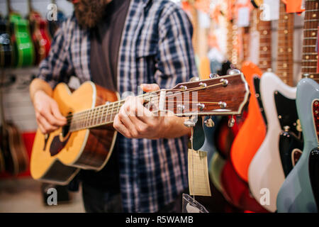 Cut vuew des Menschen stehen und Spielen auf akustischer Gitarre. Er steht im Music Shop. Viele elektronische Gitarren sind hinter Stockfoto