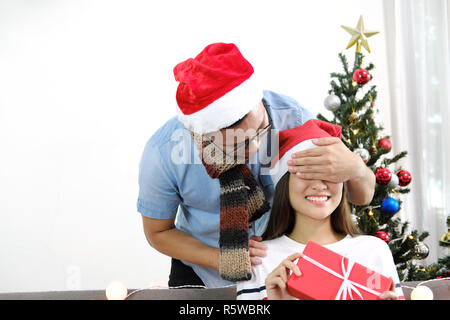 Junge Mann glücklich Überraschung schließen Freundin Augen mit den Händen mit Geschenkbox in die Weihnachtsferien. Stockfoto