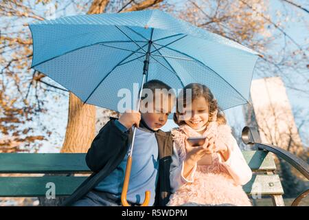 Outdoor Portrait von zwei lächelnde Kinder, Junge und Mädchen, sitzen unter einem Sonnenschirm auf der Bank im Park, am Smartphone, goldene Stunde suchen. Stockfoto