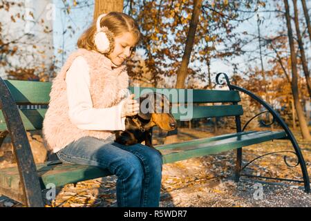 Lächelnd kleine Eigentümer eines Dackel Hund sitzt auf der Bank im Herbst Park, Mädchen mit Liebe umarmen Hund an der Goldenen Stunde. Stockfoto