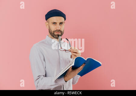 Selbstbewusst ernster Mann mit dunklen Stoppeln, nimmt Brillen, hat aufmerksamen Blick auf Kamera, hält geöffnet Lehrbuch, lernt erforderlichen Informationen, Stockfoto