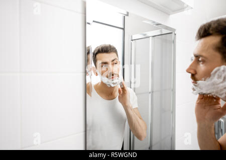 Schöner Mann im weißen T-Shirt shawing seinen Bart mit Klinge und Schaum im Badezimmer