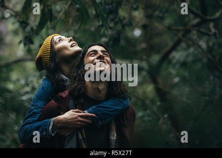 Gerne jungen Mann, seine Frau, die auf zurück in den Wald und genießen den Regen. Paar schöne im Park unter Regen huckepack. Stockfoto