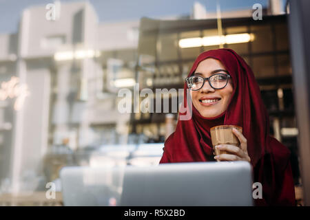 Schöne muslimische Frau mit Kopftuch und Brillen entspannen im Café eine Tasse Kaffee und Wegsehen mit schönen Lächeln auf ihrem Gesicht. Happy hija Stockfoto