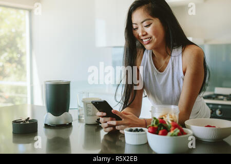 Frau lehnte den Küchentisch mit Handy am Morgen. Schöne junge Frau, die durch den Küchentisch mit Schale mit frischem Obst und readin Stockfoto