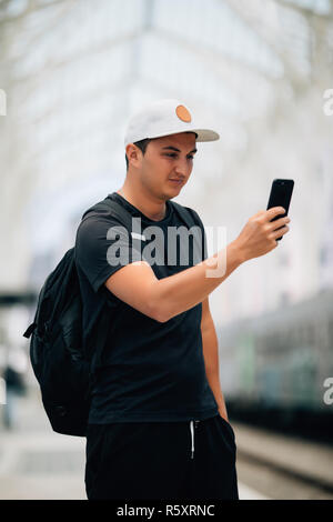 Junger Mann mit Handy mit Rucksack auf der Plattform am Bahnhof - Travel Concept, kopieren. Stockfoto