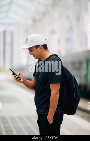 Junger Mann mit Handy mit Rucksack auf der Plattform am Bahnhof - Travel Concept, kopieren. Stockfoto