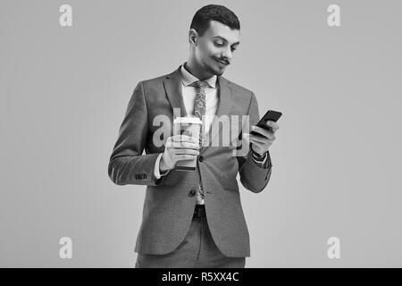 Portrait von gutaussehenden Jungen zuversichtlich arabischer Geschäftsmann mit ausgefallenen Schnurrbart in Mode grauen kompletten Anzug hält eine Tasse Kaffee und eine Meldung eingeben auf Th Stockfoto