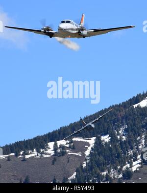 Ein U.S. Forest Service führen Ebene lässt eine Spur der weißen Rauch zu visuell ein C 130 h Luftfahrzeug verladen mit der MAFFS (Modular luftgestützten Brandbekämpfung System) von die 153 Luftbrücke Flügel der Cheyenne, Wyoming, die sich anschickt, eine Wasserleitung zu Tropfen, wie Sie trainieren wildfires außerhalb Boise, Idaho zu enthalten. April 21, 2017. Mehr als 400 Mitarbeiter von vier C-130 Schutzes und der Reserve Einheiten - von Kalifornien, Colorado, Nevada und Utah, der Air Expeditionary Gruppe - sind in Boise, Idaho für die Woche - lange wildfire Schulung und Zertifizierung durch die US-gesponserten Stockfoto