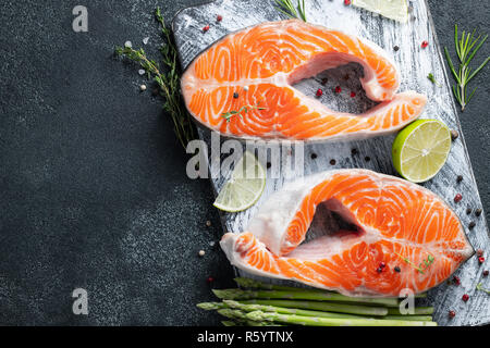 Zwei rohe frische Lachs oder Forelle Steaks auf Eis, die reich an Omega-3-Öl, mit Kalk, Thymian und junge Spargel auf einem dunklen Hintergrund. Gesunde und diätetische Lebensmittel. Stockfoto