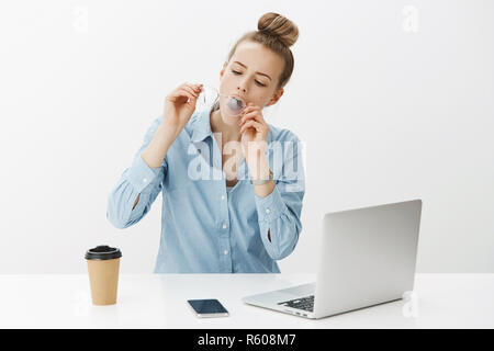 Mädchen die Atmung auf Gläser und Reinigung das Objektiv vor Staub, whiping Eyewear, als faul nicht arbeiten wie im Büro in der Nähe von Laptop und Smartphone, Abakus Stockfoto