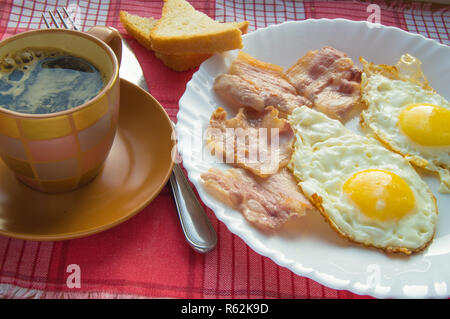 Köstliches Frühstück - eine Tasse Kaffee, einen Teller mit Spiegelei, Speck und Toast, neben dem Besteck auf rot kariert Serviette Stockfoto