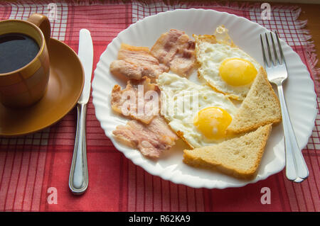 Köstliches Frühstück - eine Tasse Kaffee, einen Teller mit Spiegelei, Speck und Toast, neben dem Besteck auf rot kariert Serviette Stockfoto