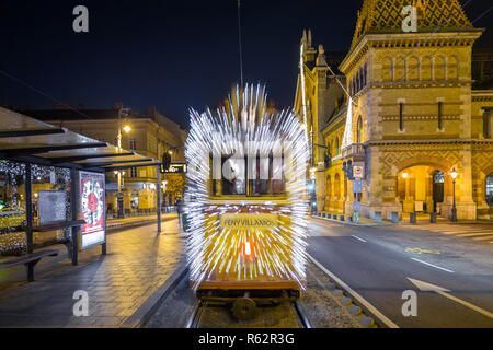 Budapest, Ungarn - festlich geschmückten Licht Straßenbahn (Fenyvillamos) unterwegs an Fovam Platz mit der Großen Markthalle (Vasarcsarnok) bei Nacht. Chri Stockfoto