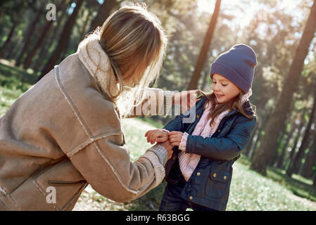 Junge Mutter hilft, Ihr kleines lockiges Mädchen zu Mantel, während Sie zu Fuß in den Wäldern. Kalte Jahreszeit, helle Sonne ist, die durch die Bäume im Wald gesehen Stockfoto