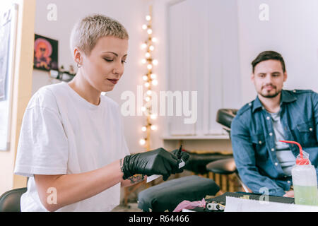 Kurzhaarige blonde Frau mit schwarzen Handschuhen und Vorbereitung Tinte Maschine Stockfoto