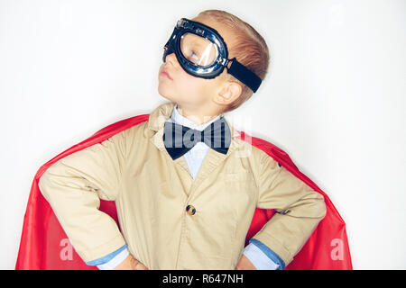 Adorable kleinen Jungen Superhelden in Anzug und Fliege tragen Schutzbrille und einem roten Cape suchen mutige während vor einem weißen Hintergrund stehen Stockfoto