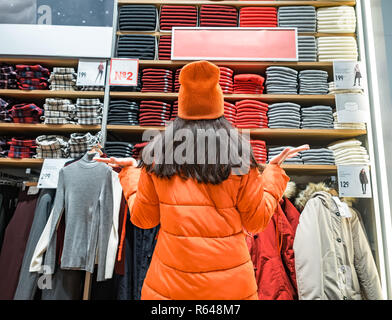 Ansicht der Rückseite des verwirrten Frau, die Gestik, die Hände zur Seite, während sie Kleidung in Store angezeigt. Viele warme helle farbige Pullover verschiedener Farbe sind ordentlich auf dem Regal gestapelt Stockfoto