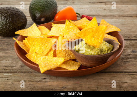 Holzplatte mit Nachos und Guacamole, Avocados, Pfeffer und Chili im Hintergrund auf rustikalen Tisch Stockfoto