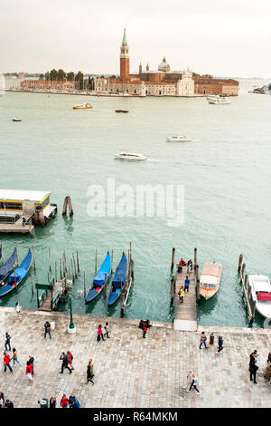 Blick auf Saint Giorgio Insel aus der Distanz über Wasser mit Booten. Touristen wandern auf dem Deich am Hafen im Vordergrund. Stockfoto