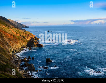 Die untergehende Sonne beleuchtet die Rot-, Grün- und Brauntönen der steile Berge und Klippen unter einem blauen Himmel entlang der Kalifornien Big Sur Küste Stockfoto