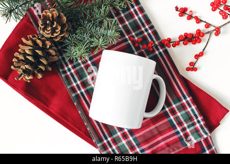 Winter noch leben. Leere Kaffeetasse, Fir Tree Branch, Stechpalme Beeren und Tannenzapfen. Kariert tartan plaid. Weihnachten im traditionellen Stil Zusammensetzung, rustikalen Szene. Produkt mockup. Flach, Ansicht von oben. Stockfoto