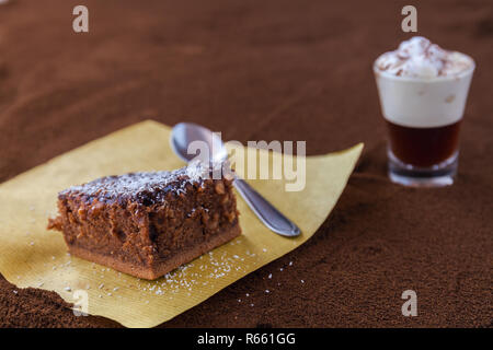 Kleine Tasse piccolo Latte macchiato auf einem Tisch mit gemahlenen Kaffee als Hintergrund und Chocolate Brownie mit Kakao und Sahne coconut Chips Stockfoto