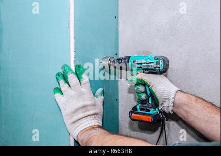 Ein Arbeiter bohrt Löcher in einem gipskartonplatten Wand mit einer elektrischen Bohrmaschine. Reparatur im Haus. Stockfoto