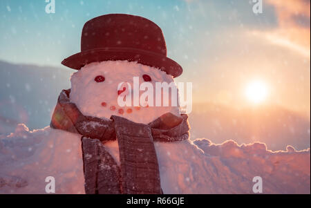 Foto von einem niedlichen Schneemann in einem stilvollen Hut und Schal in einem schneebedeckten eisigen Abend an einem Sonnenuntergang Hintergrund gekleidet, glückliche Winterferien tradition Stockfoto