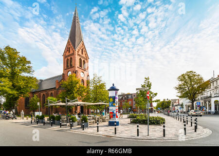 Die neugotischen Warnemünder Kirche Turm erhebt sich über die Altstadt von Rostock, Deutschland, als Touristen und Deutsche speisen Sie auf der Terrasse im Freien Platz. Stockfoto