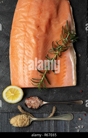 Roher Lachs filet auf schwarzem Schiefer Platte und Zutaten für die Herstellung von Graved Lachs. Salz, Zucker, Zitrone und Rosmarin im Hintergrund für trockene Heilung Marinade, da Stockfoto