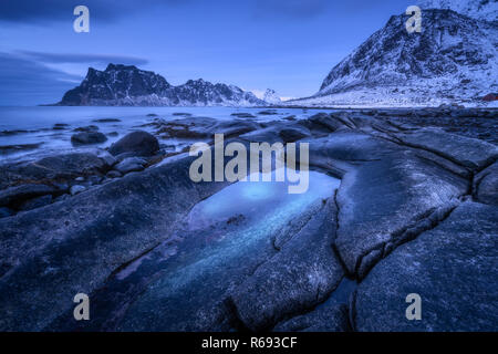 Seashore mit Steinen und fließend Wasser, gegen verschneite Berge und blauer Himmel mit Wolken bei Sonnenuntergang. Uttakleiv Strand auf den Lofoten Inseln, Norwegen. Winter l Stockfoto