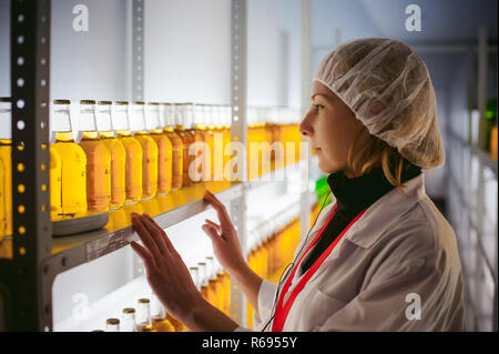 Mitarbeiter der Bierherstellung Labor. Junge Lab Assistant, weißes Gewand gekleidet, steht auf Bier Lagerverwaltung. im Hintergrund sind leichte Regale mit Bierflaschen. Bier-Abteilung Forschung Stockfoto