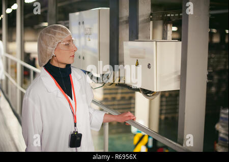 Arbeiter in der Nähe von elektrischen Abschirmung. Eine Frau in einem weißen Gewand und Schutzbrille, hält sich an die Rampe, in der Nähe der Schalttafel. In der Fertigung im Innenbereich. Arbeitsschutz auf die Produktion Stockfoto