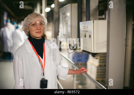 Arbeiter in der Nähe von elektrischen Abschirmung. Eine Frau in einem weißen Gewand und Schutzbrille, hält sich an die Rampe, in der Nähe der Schalttafel. In der Fertigung im Innenbereich. Arbeitsschutz auf die Produktion Stockfoto