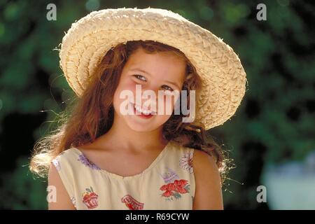 Portrait, Outdoor, laechelndes Maedchen mit langen braunen Haaren und braunen Augen, 6 Jahre alt, bekleidet mit buntem Sommerkleid und Strohhut Stockfoto