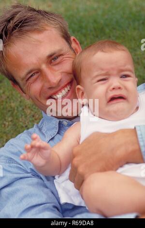 Outdoor, Portrait, lachender Vater im Jeanshemd mit seinem weinenden Baby im Arm in einer Wiese Stockfoto