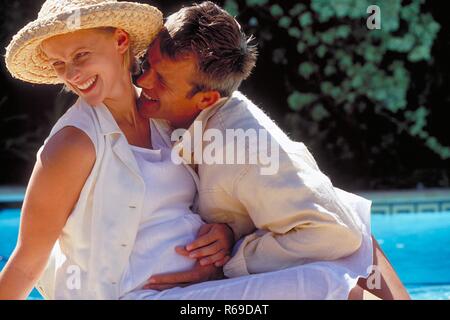 Outdoor, Profil, blonder Mann moechte seine schwangere weiss gekleidete Frau mit Strohhut am Rand eines Schwimmbeckens kuessen Stockfoto