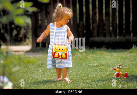 Kleines blondes Maedchen mit blau-weiss gestreiftem Kleid und Umhaengetasche spielt mit Zugente im Garten Stockfoto