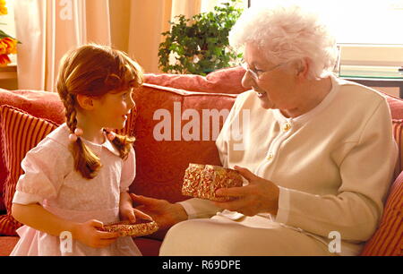 Porträt, Grossmutter sitzt auf dem Sofa und packt mit ihrer Enkeltochter ein Geschenk aus Stockfoto