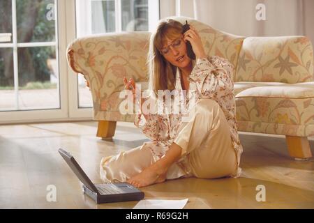 Innenraum, Porträt, Ganzfigur, Frau Mitte 30 blondn mit langen Haaren und Brille sitzt im Schlafanzug mit ihrem Laptop vor dem Sofa auf dem Fussboden und telefoniert Stockfoto