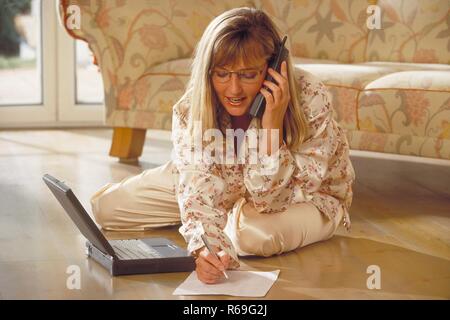 Innenraum, Porträt, Ganzfigur, Frau Mitte 30 blondn mit langen Haaren und Brille sitzt im Schlafanzug mit ihrem Laptop vor dem Sofa auf dem Fussboden, telefoniert und macht sich Notizen Stockfoto