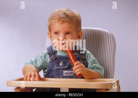 Porträt, Innenraum, Ca. 1-jaehriger blonder Junge bekleidet mit Jeanslatzhose sitzt mit einem wuerstchen in der Hand in einem Hochstuhl Stockfoto